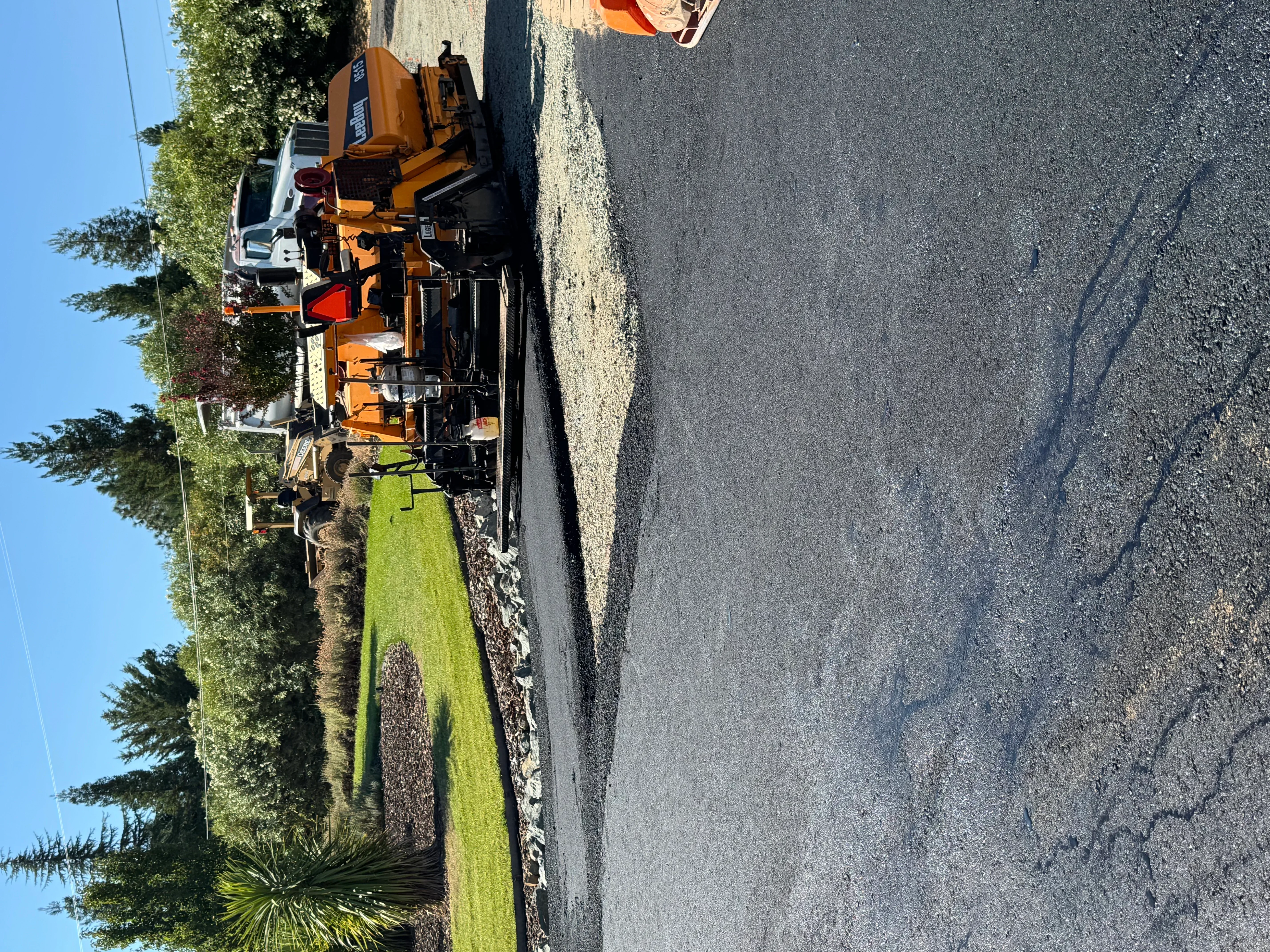 Orange paving truck laying fresh asphalt on residential driveway with green lawn and trees